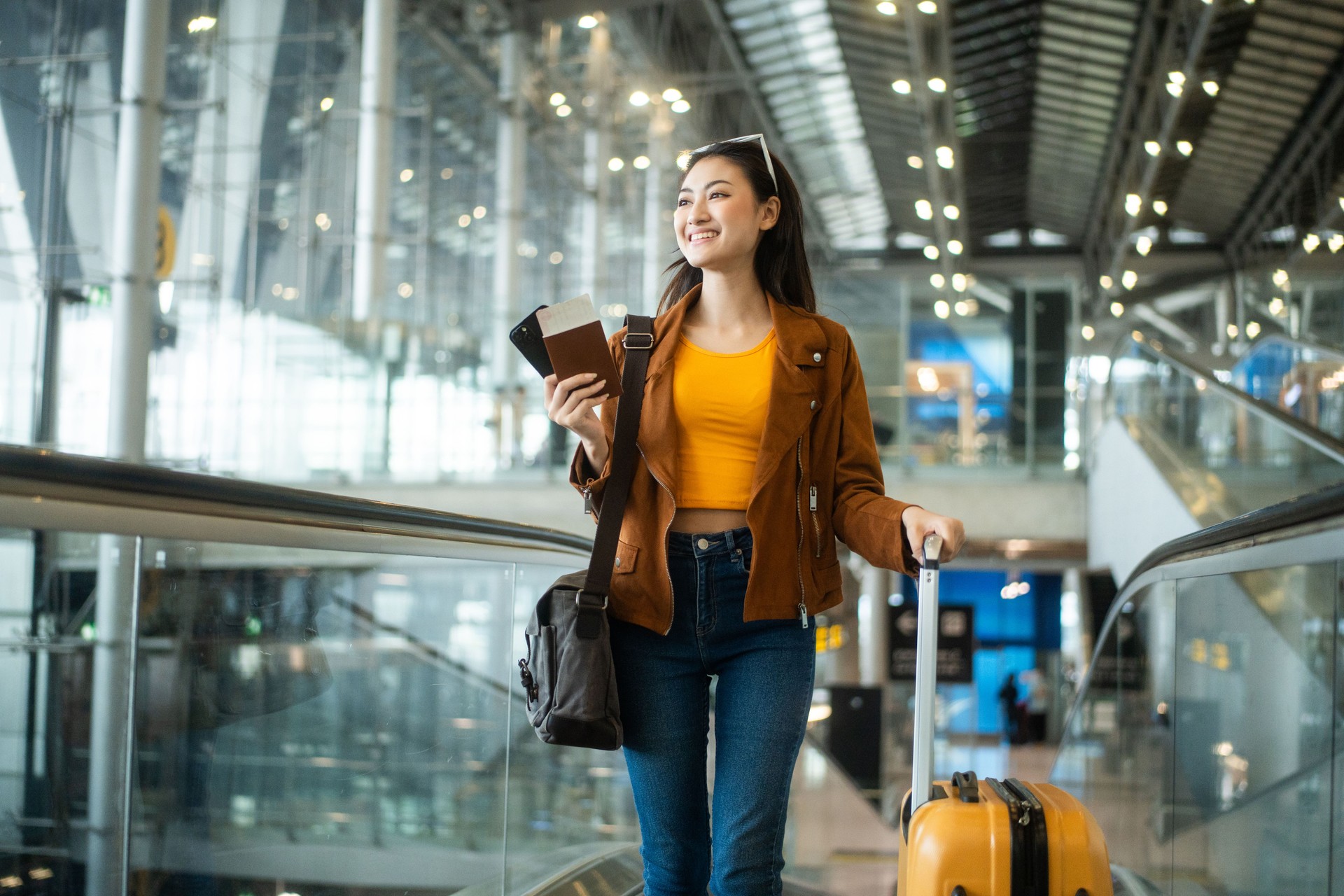 happy young asian woman traveler holding boarding pass ticket, passport and smartphone at the airport terminal with her luggage and smiling at camera, cheerful tourist female having holiday trip.